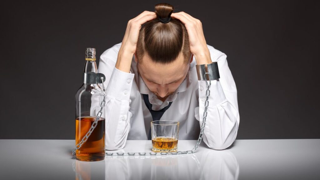 Alcohol Detox vs. Drug Detox: What’s the Difference? 2 A young man in a white shirt holding his head with both palms, looking down, while his left arm is chained to a bottle of hard liquor.