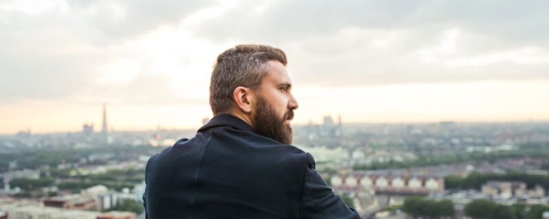A man rests on a rooftop deck of a corporate skyscraper at sunset, surrounded by city buildings, reflecting on recovering without quitting his career.
