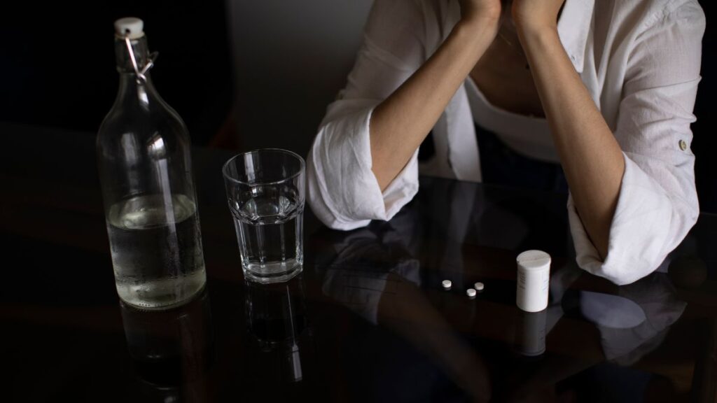 5 Signs You’re In Denial About Substance Use Disorder 2 Denial About Substance Use Disorder: A man rests his elbow on a table scattered with bottles of hard liquor and pills, only his torso visible.