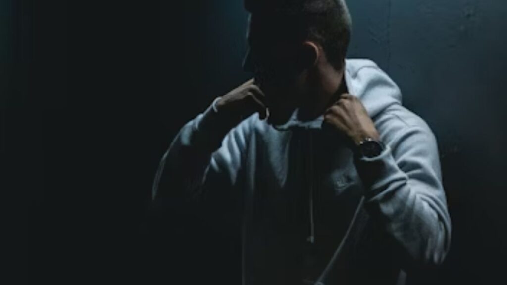 The Hidden Crisis: Professionals Hiding Addiction 3 A young man in a hooded shirt walks through a dark alley and pauses against a wall, his face and body hidden in shadow, symbolizing professionals hiding addiction.