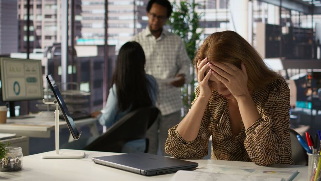 How Professionals Can Overcome Addiction While Maintaining Their Careers 2 A frustrated female professional rests her elbows on a desk, covering the sides of her forehead with her hands, while coworkers chat in the blurred background.